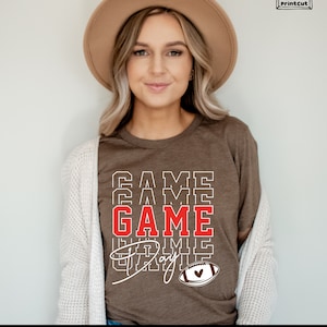 May include: A brown t-shirt with the text "GAME DAY" in white and red letters. The text is stacked on top of itself with the word "GAME" in red. A brown and white football with a red heart is below the text.