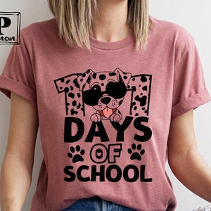 K&ouml;nnte beinhalten: Ein pinkfarbenes T-Shirt mit einem schwarz-wei&szlig;en Dalmatiner, der eine Sonnenbrille tr&auml;gt und den Text "101 Tage Schule" mit Pfotenabdr&uuml;cken und einem Herzen.