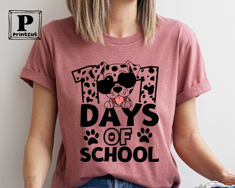K&ouml;nnte beinhalten: Ein pinkfarbenes T-Shirt mit einem schwarz-wei&szlig;en Dalmatiner, der eine Sonnenbrille tr&auml;gt und den Text "101 Tage Schule" mit Pfotenabdr&uuml;cken und einem Herzen.
