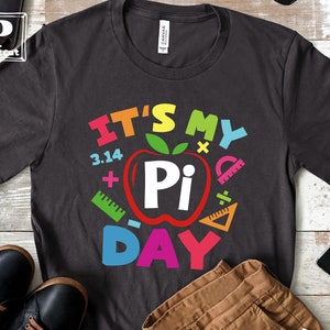 May include: A dark gray t-shirt with the text "It's My Pi Day" in colorful letters. The text is surrounded by colorful math symbols, including a red apple with a white "Pi" inside.