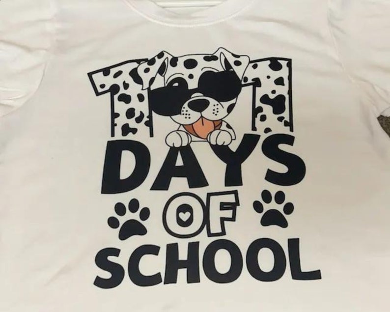 K&ouml;nnte beinhalten: Wei&szlig;es T-Shirt mit einem schwarz-wei&szlig;en Dalmatiner, der eine Sonnenbrille tr&auml;gt und den Text "101 Tage Schule" mit Pfotenabdr&uuml;cken.