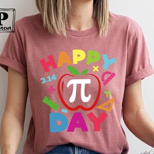 May include: A pink t-shirt with the text "Happy Pi Day" in rainbow colors. The design includes a red apple with a white pi symbol and other math-related symbols.