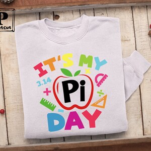 May include: White sweatshirt with a colorful graphic design that says "It's My Pi Day" with a red apple with a black "Pi" inside and other colorful math symbols.