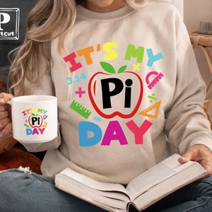 May include: A white ceramic mug with a colorful graphic that says "It's My Pi Day" with a red apple and the word "Pi" inside. The mug is being held by a person wearing a beige sweatshirt with the same graphic.