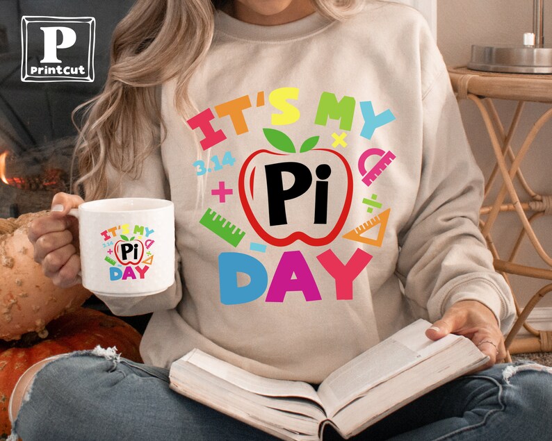 May include: A white ceramic mug with a colorful graphic that says "It's My Pi Day" with a red apple and the word "Pi" inside. The mug is being held by a person wearing a beige sweatshirt with the same graphic.