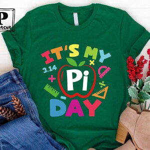 May include: Green t-shirt with the text "It's My Pi Day" in rainbow colors. The text is surrounded by a red apple with a white "Pi" inside and math symbols.