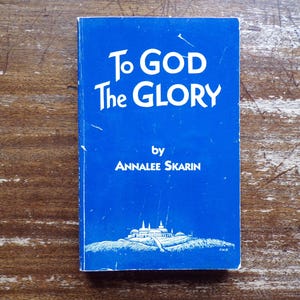 May include: A small, blue book titled "To GOD The GLORY" by Annalee Skarin. The book cover features white text and a small illustration of a building. The book is set against a wooden background.