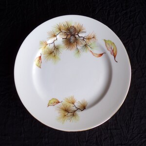 May include: White porcelain plate with a gold rim and a painted design of pine branches and cones in shades of green, brown, and yellow.