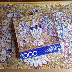 May include: A 1000-piece jigsaw puzzle featuring a gold eagle with a shield that says "The Authentic All-American Bicentennial Jigsaw Puzzle". The eagle is surrounded by a variety of small images, including stars, flags, and other patriotic symbols.