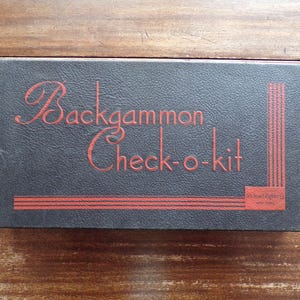 May include: A vintage backgammon check-o-kit box. The box is dark gray with red lettering and design elements. The words "Backgammon Check-o-kit" are prominently displayed in a decorative font. The box has a rectangular shape.