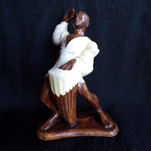 May include: A ceramic figurine of a person in a dynamic pose, with one arm raised. The figure has dark skin and is wearing a white outfit with a cream-colored, scalloped edge. The base and lower portion are brown.
