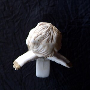 May include: A small, cream-coloured figurine of a person in a seated position, with arms outstretched. The figure has detailed hair and is wearing a long-sleeved garment. The sculpture is set against a dark background.