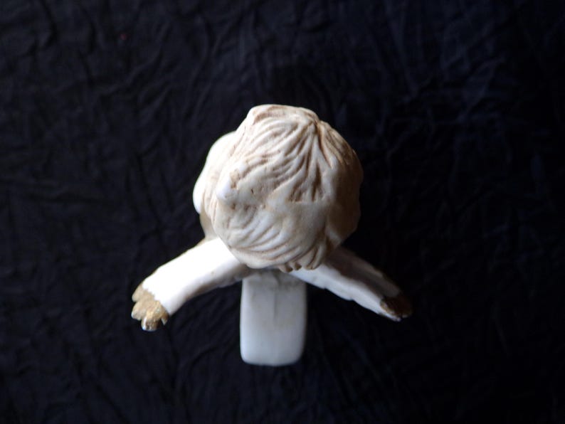 May include: A small, cream-coloured figurine of a person in a seated position, with arms outstretched. The figure has detailed hair and is wearing a long-sleeved garment. The sculpture is set against a dark background.