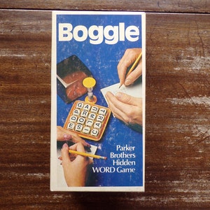 May include: A blue and white box for the Boggle word game. The box shows a close-up of the game board with letter tiles and a hand writing on a piece of paper. The text on the box reads "Boggle" and "Parker Brothers Hidden Word Game".