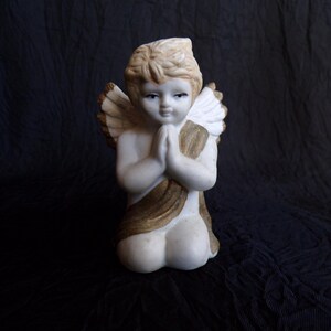 May include: A kneeling angel figurine with folded hands, featuring a cream-coloured body, blonde hair, and detailed wings. The angel is adorned with a gold-coloured sash. The statue is set against a dark background.