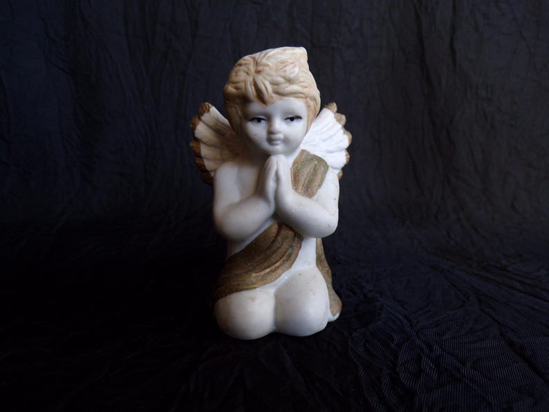 May include: A kneeling angel figurine with folded hands, featuring a cream-coloured body, blonde hair, and detailed wings. The angel is adorned with a gold-coloured sash. The statue is set against a dark background.