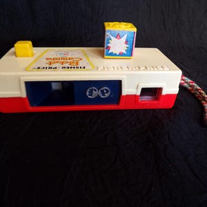 May include: Vintage Fisher-Price Pocket Camera toy. The camera is primarily off-white with red and blue accents. A small yellow square and a colorful block with a starburst design sit on top. A multi-colored cord is attached to the side.