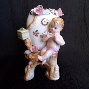 May include: A porcelain figurine of a cherub seated on a tree branch, holding a flute. The figurine is part of a vase, decorated with pink roses and floral accents. The cherub wears a pink cloth. The vase has a small green box on the side.