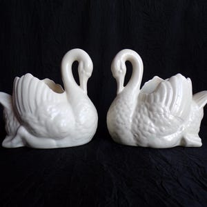 May include: Two off-white ceramic swan planters. Each swan has a curved neck and open top for plants. The wings are detailed with textured feathers. The swans are positioned facing each other against a dark background.