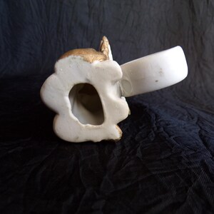 May include: A white ceramic angel figurine with gold detailing. The angel is holding a white cup. The figurine has a detailed face and wings. It is set against a dark background.