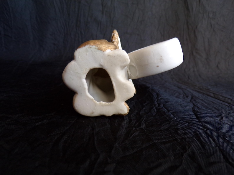 May include: A white ceramic angel figurine with gold detailing. The angel is holding a white cup. The figurine has a detailed face and wings. It is set against a dark background.