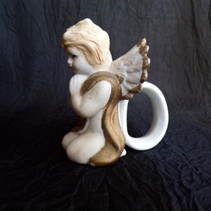 May include: A ceramic angel figurine in a kneeling pose, with a white body, blonde hair, and gold detailing on the wings and clothing. The angel has a white handle attached to the back, creating a unique design.