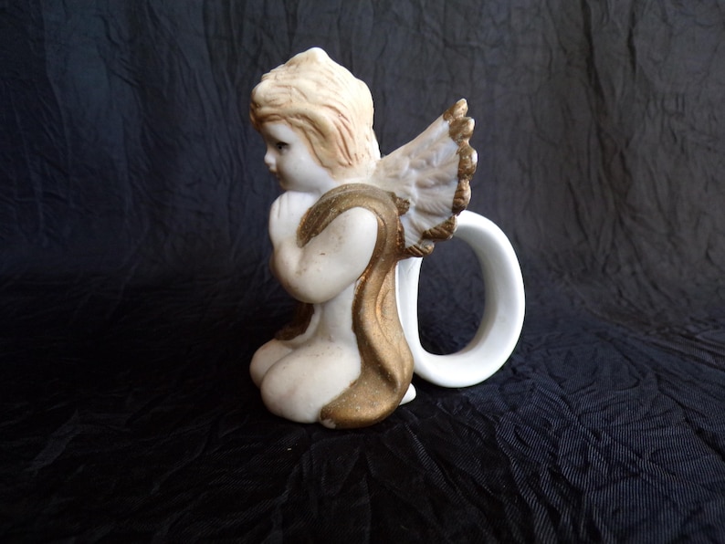 May include: A ceramic angel figurine in a kneeling pose, with a white body, blonde hair, and gold detailing on the wings and clothing. The angel has a white handle attached to the back, creating a unique design.