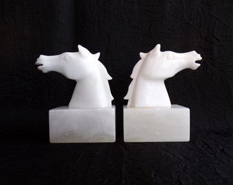 May include: A pair of white alabaster bookends shaped like horse heads. The bookends are on white bases.