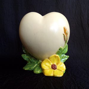 May include: Cream-colored heart-shaped vase with a yellow flower and green leaves at the base. The flower has a dark red center. A small, brown and green bud is on the right side of the heart. Decorative ceramic vase.