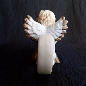 May include: A ceramic angel figurine, viewed from the back, with detailed white wings edged in brown. The angel has light brown hair and is holding a cream-coloured object. The figurine is set against a dark background.
