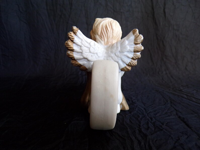 May include: A ceramic angel figurine, viewed from the back, with detailed white wings edged in brown. The angel has light brown hair and is holding a cream-coloured object. The figurine is set against a dark background.