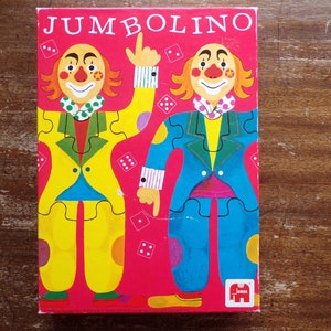 May include: A red box with the word "JUMBOLINO" in white letters. The box features two clowns with colorful costumes and puzzle pieces. The clowns are wearing yellow and blue suits with red shoes. The box has a small elephant logo in the bottom right corner.