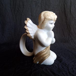 May include: A white ceramic angel figurine with a golden sash and wings. The angel is kneeling with hands clasped in prayer. The wings have a detailed feather design. The figurine has a ring-shaped handle.