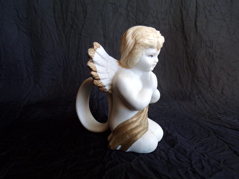 May include: A white ceramic angel figurine with a golden sash and wings. The angel is kneeling with hands clasped in prayer. The wings have a detailed feather design. The figurine has a ring-shaped handle.