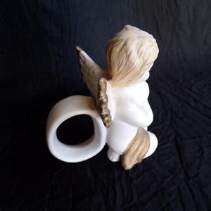 May include: A ceramic angel figurine in a kneeling pose, with a white body, tan hair, and gold-accented wings. The angel is holding a white ring, possibly a napkin ring, against a dark background.
