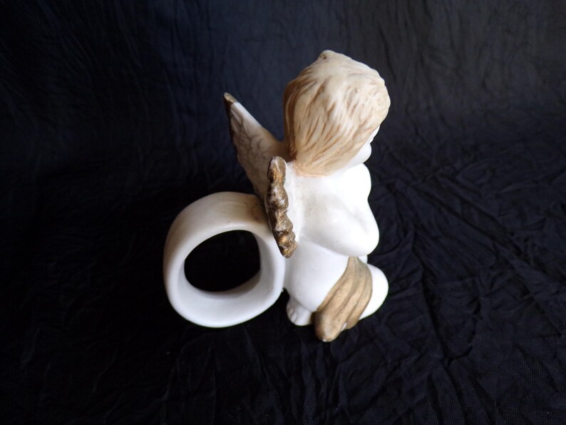 May include: A ceramic angel figurine in a kneeling pose, with a white body, tan hair, and gold-accented wings. The angel is holding a white ring, possibly a napkin ring, against a dark background.