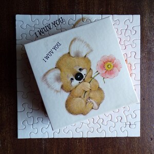 May include: A square puzzle with a box featuring a koala holding a pink flower. The box reads "I WUV YOU." The puzzle pieces are white, and the box is on top of the assembled puzzle.