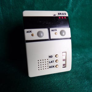 May include: A rectangular, off-white electronic device with black and gray accents. It features buttons labeled "ACK" and "NU15", along with the text "ER-2/3". The device has a series of small holes and indicator lights.