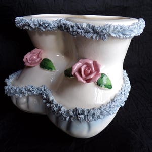 May include: A white ceramic vase with a unique shape, adorned with pink rose accents and light blue trim. The vase has a ruffled edge and a glossy finish, creating a vintage aesthetic. The vase is a decorative item.