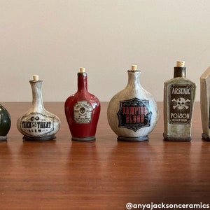 May include: A set of seven small ceramic bottles with cork stoppers and Halloween-themed labels. The labels include "Trick or Treat Brew", "Vampire Blood", "Arsenic Poison", and a skull and crossbones.