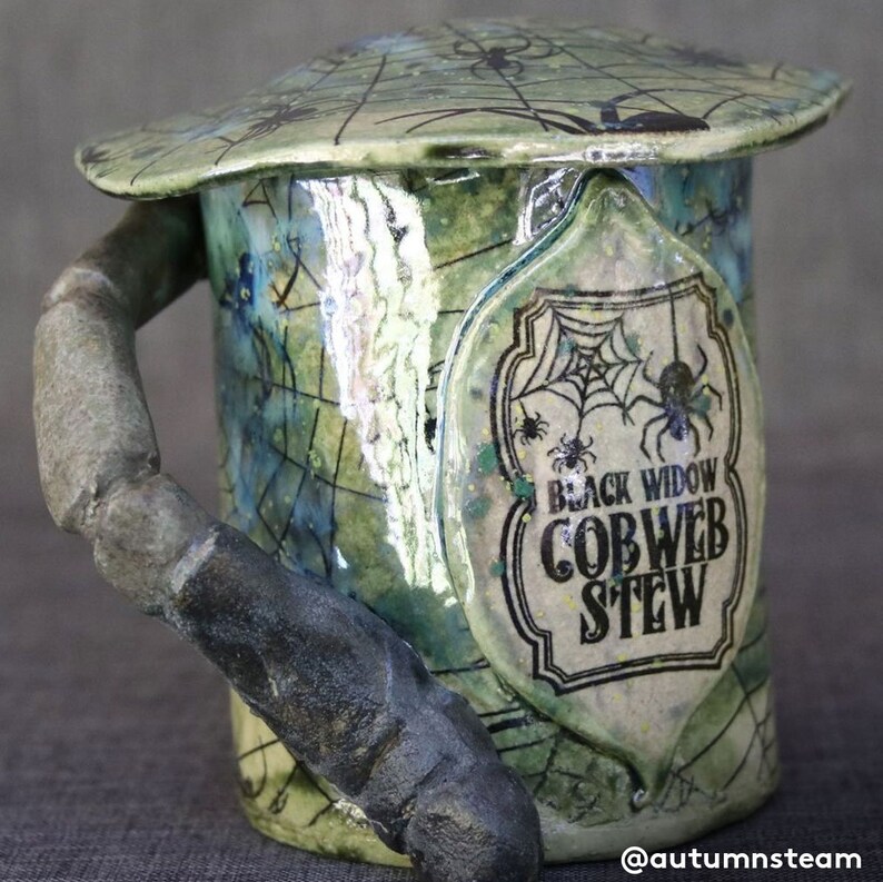 May include: A green ceramic mug with a spiderweb design and a black label that reads "Black Widow Cobweb Stew". The mug has a unique, textured handle shaped like a spider leg.