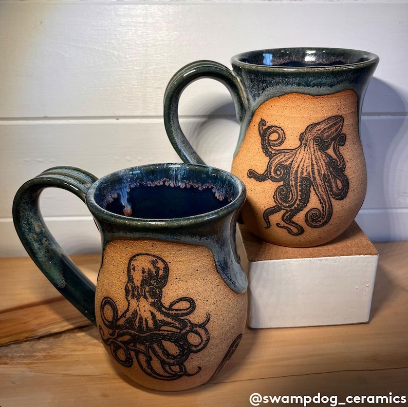 Op de afbeelding: Twee keramische mokken met een donkerblauwe glazuur en een lichtbruin interieur. Elke mok heeft een zwart-witte illustratie van een octopus.