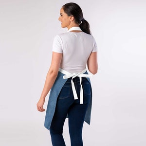 May include: A blue denim apron with white straps tied at the back. The apron is worn over a white t-shirt and blue jeans.