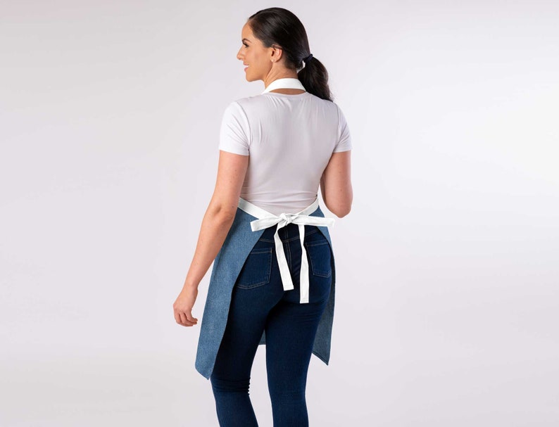 May include: A blue denim apron with white straps tied at the back. The apron is worn over a white t-shirt and blue jeans.