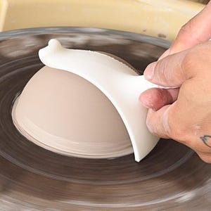 May include: A potter's hands shaping a light brown ceramic bowl on a spinning pottery wheel. A white tool is used to refine the bowl's form. The wheel and bowl are in motion, creating a dynamic scene.