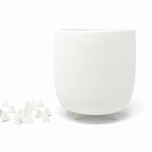以下が含まれることがあります： 丸みを帯びた形をした白いセラミック製のプランターで、小さな三角形の足が3つ付いています。プランターは白い表面の上に置かれており、その前に小さな白い三角形のものがいくつか散らばっています。