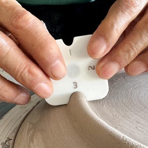 May include: A white pottery tool marked with the numbers 1, 2, and 3 is used to shape clay. The tool is held by two hands, and the clay sits on a pottery wheel. The tool is used to measure the thickness of the clay in centimetres.