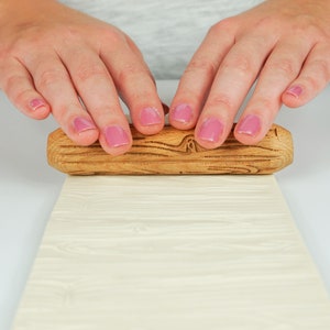 May include: A wooden rolling pin with a textured surface is being used to roll out a sheet of white fondant. The rolling pin has a natural wood finish and is being held by two hands with pink nail polish.