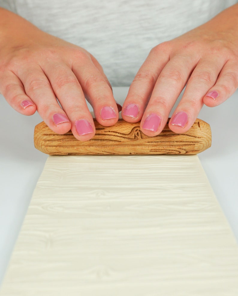 May include: A wooden rolling pin with a textured surface is being used to roll out a sheet of white fondant. The rolling pin has a natural wood finish and is being held by two hands with pink nail polish.
