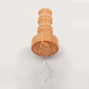 May include: A wooden stamp with a leaf design. The stamp is used to create an impression of a leaf on a surface.